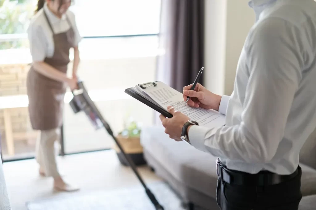 An employer taking a checklist, his transfer domestic helper vacuums the carpet in the background.