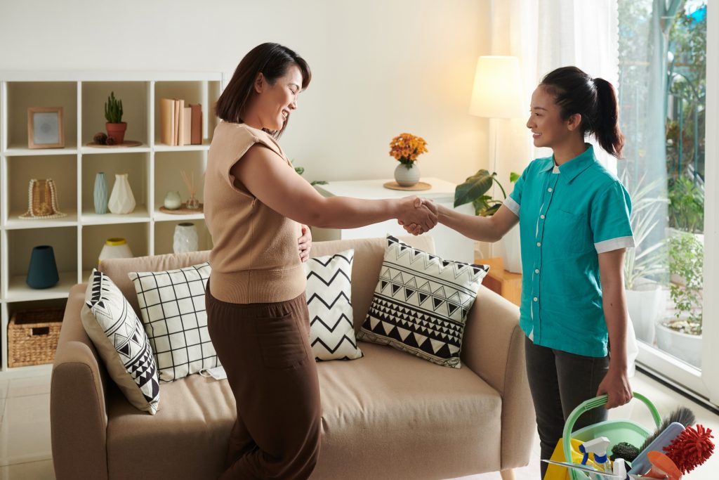 In the living room, an employer shakes her FDW's hand, indicating rapport and a good working relationship. The FDW holds cleaning supplies in the other hand.